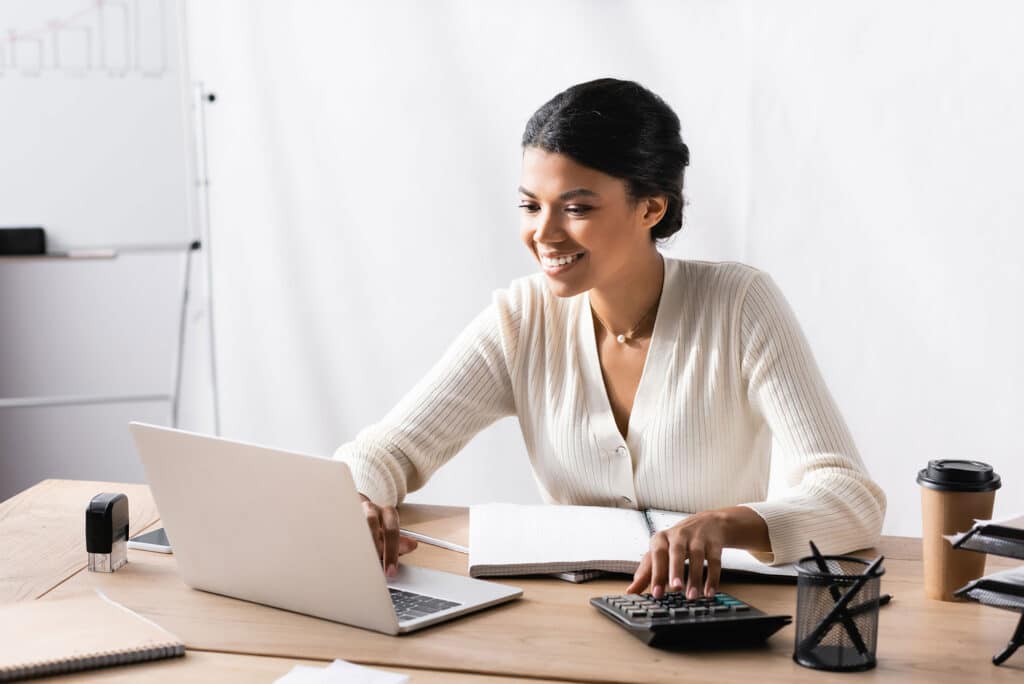 Happy african american woman looking at laptop while accounting on calculator in office on blurred background