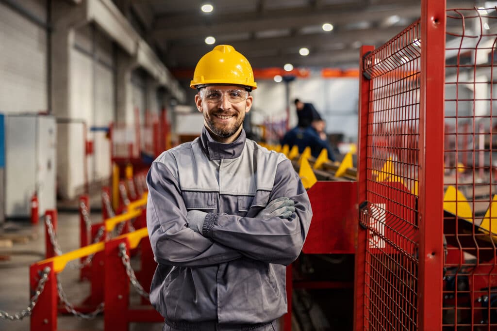 A heavy industry worker stands in a facility with arms crossed and smiling at the camera.